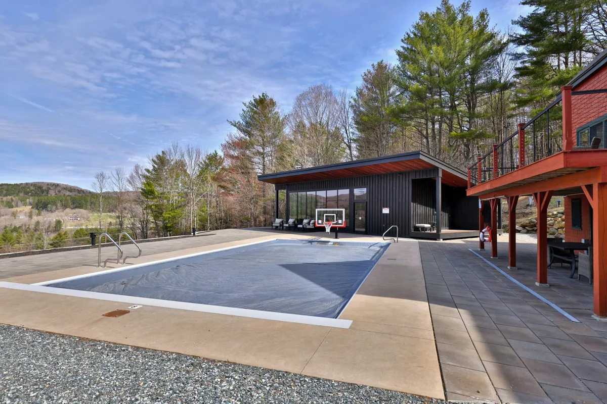 Close-up of the swimming pool featuring the basketball hoop, modern pool house, and tall trees that provide both privacy and a natural resort-like atmosphere. The space is set up for sunbathing, swimming, and active play.