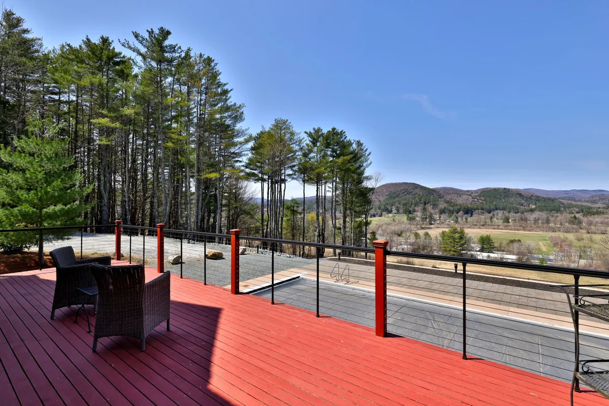 A tranquil red wooden deck furnished with wicker chairs, overlooking lush green trees and distant mountains. The inviting setup is ideal for sunset cocktails, casual gatherings, or quiet moments appreciating the view.