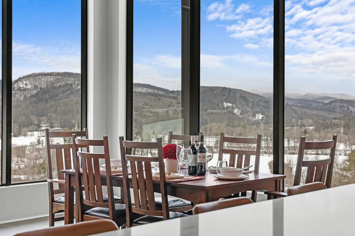 An interior view focused on a dining table set near large windows that frame panoramic mountain views. The table is set for an intimate meal, with wine bottles and a cozy atmosphere suggesting special celebrations or quiet dinners. The natural light and vista create a sense of connection with the outdoors even while inside.