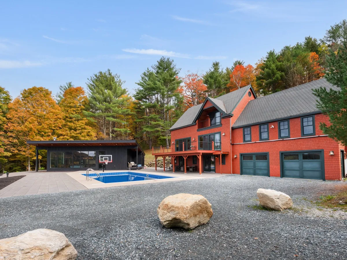 A striking exterior shot featuring a modern red house, a heated swimming pool, black outdoor structure, and colorful autumn trees. The scene is lively, showcasing the pool as the centerpiece surrounded by space for barbecuing, basketball, and relaxation, all set against a backdrop of seasonal foliage.