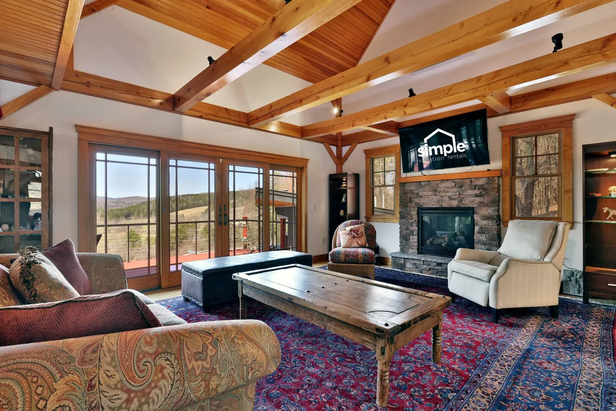 A cozy living room centered around a rustic stone fireplace, with exposed wooden beams, large windows, and plush seating inviting guests to gather and unwind. The space exudes warmth and offers glimpses of the natural landscape outside.