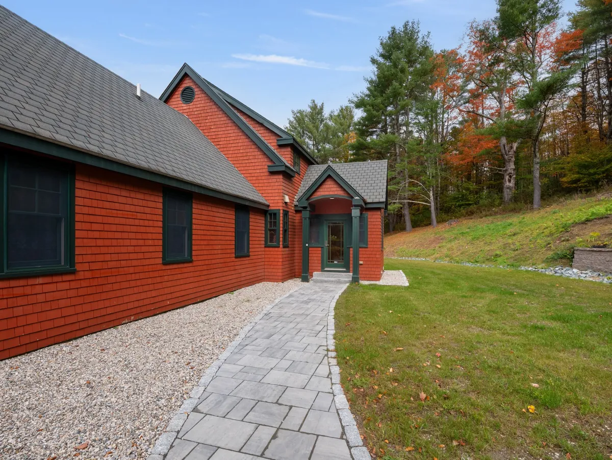 The red exterior of the house, warmly accented by green-trimmed windows and a welcoming front pathway, is surrounded by vibrant greenery. The inviting entrance hints at easy access to both privacy and local attractions.