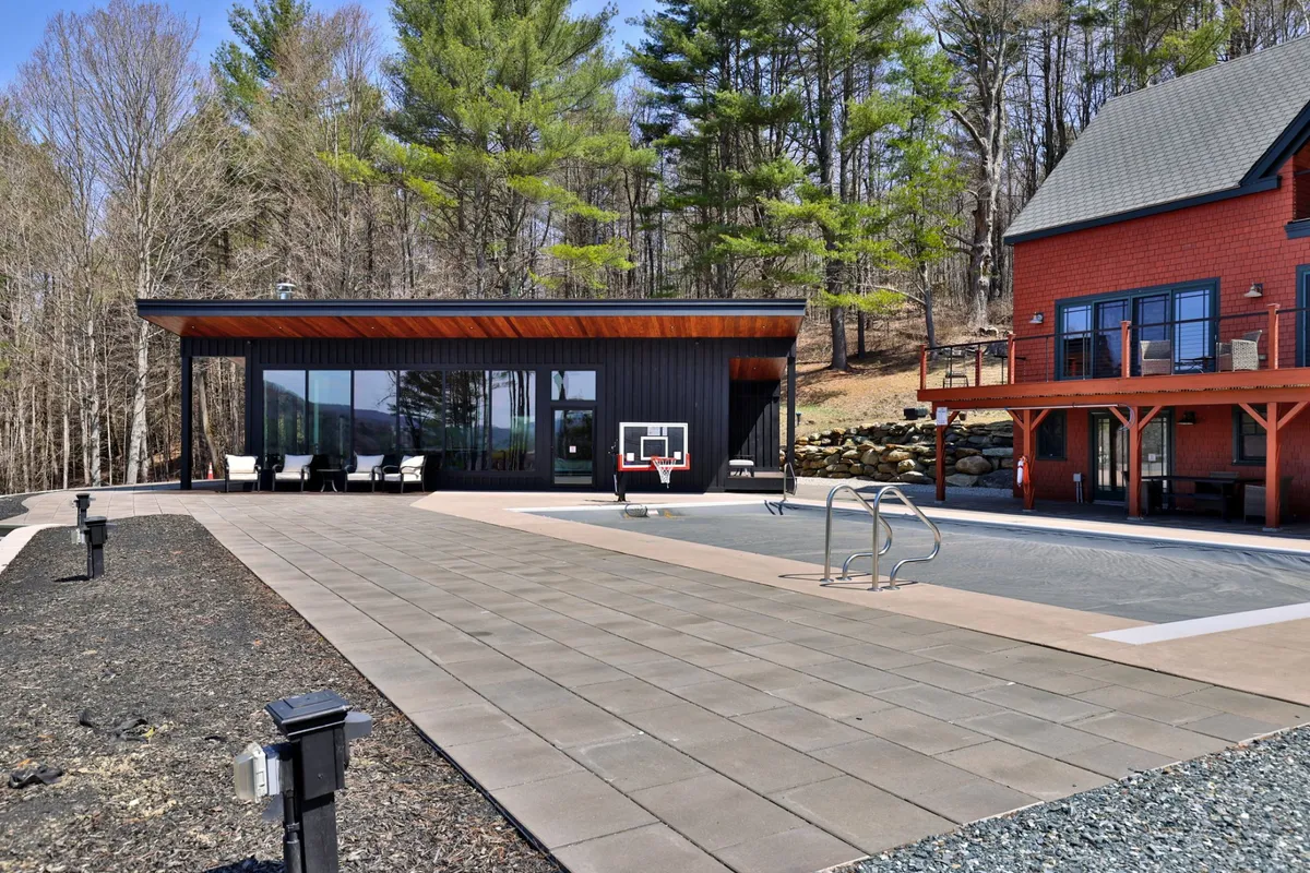 This image features the modern black pool house, a sparkling swimming pool, and a poolside basketball hoop. The wooden deck and relaxed seating reinforce the sense of a private resort space, while surrounding greenery ensures privacy and a tranquil backdrop.