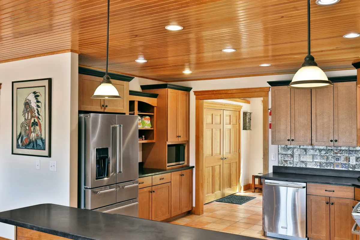 This image centers on the main house kitchen with a prominent kitchen island, high-end refrigerator, custom wood cabinetry, pendant lighting, and tasteful wall artwork. The mood is inviting and warm, with visual cues of cooking, entertaining, and family gatherings. Granite countertops and the overall spacious design suggest an ideal environment for both everyday meals and special celebrations.