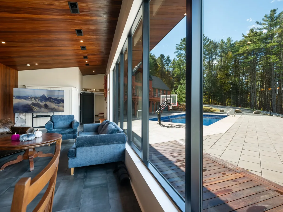 An inviting interior lounge area with large windows overlooking the pool, plush seating, and a warm wooden ceiling. This space blends seamless indoor-outdoor living, with radiant heated floors likely present and a calming sunset ambiance visible through the glass.